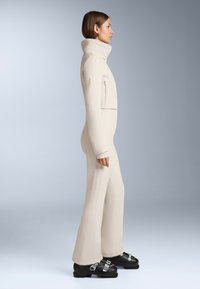 Beżowy kombinezon pełnej długości z wysokim kołnierzem i zapięciem z przodu, charakteryzujący się gładkim, dopasowanym krojem oraz subtelnym detalem szwów, zestawiony z czarnym obuwiem.