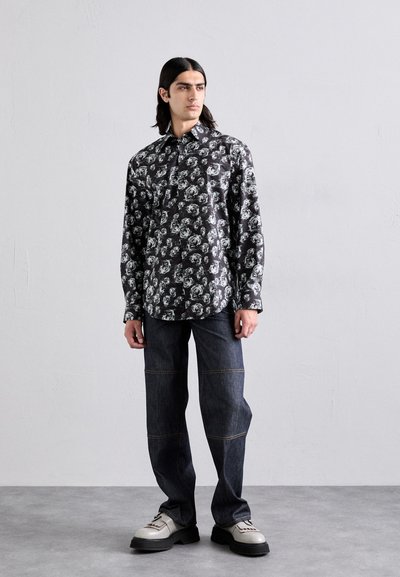Black shirt with white floral pattern, long sleeves, buttoned collar; paired with dark jeans featuring contrasting stitching and chunky shoes.