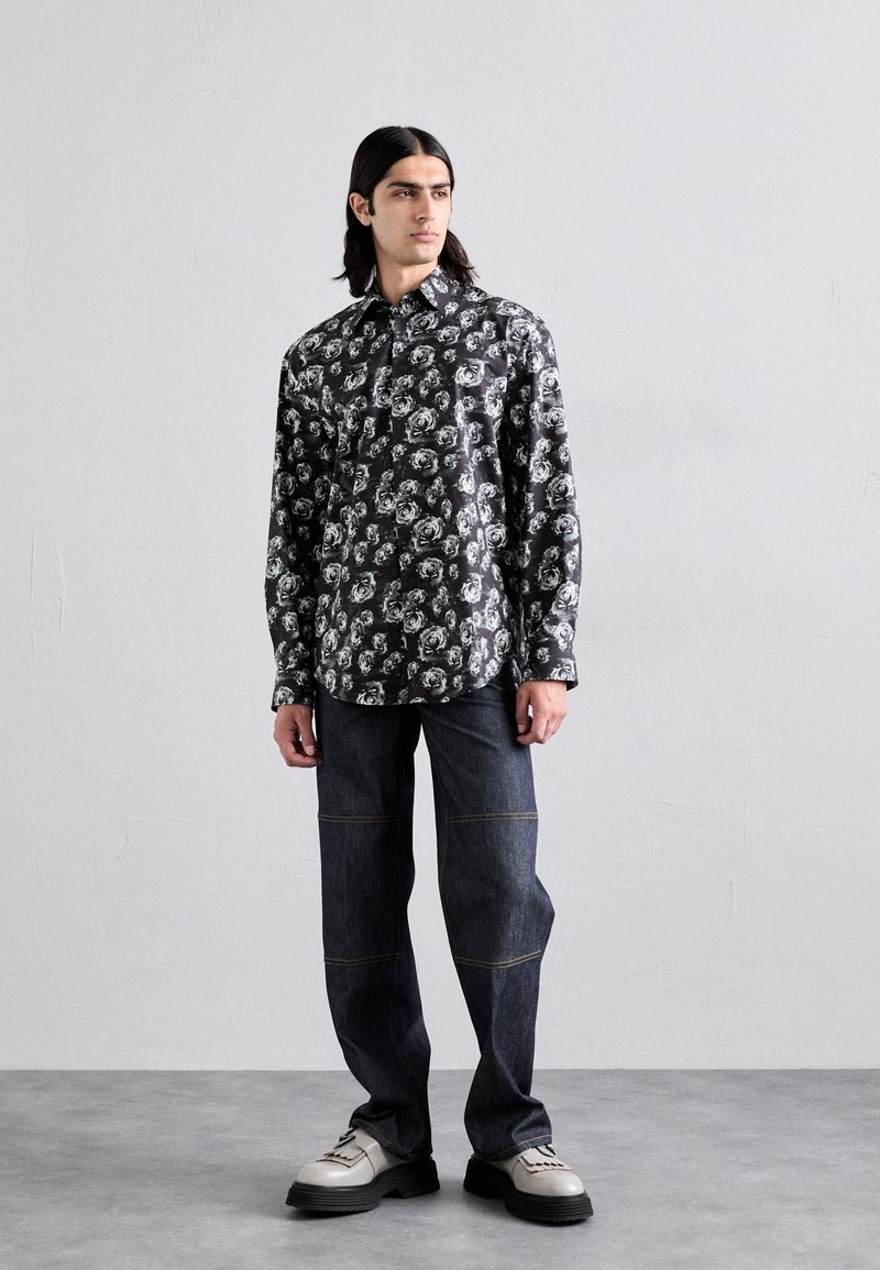 Schwarzes Hemd mit weißem Blumenmuster, lange Ärmel, geknöpfter Kragen; kombiniert mit dunklen Jeans mit kontrastierenden Nähten und klobigen Schuhen.