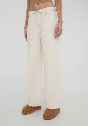 Jeans a gamba larga di colore crema realizzati in tessuto resistente, con chiusura anteriore a legaccio e tasche. Abbinati a scarpe slip-on di colore marrone chiaro.