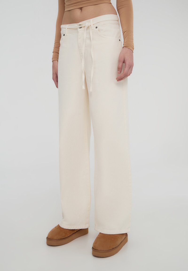 Jeans a gamba larga di colore crema realizzati in tessuto resistente, con chiusura anteriore a legaccio e tasche. Abbinati a scarpe slip-on di colore marrone chiaro.