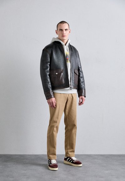 Veste bombardier en cuir marron avec fermeture éclair et deux poches avant, portée sur un hoodie gris, associée à un pantalon beige et des baskets multicolores.