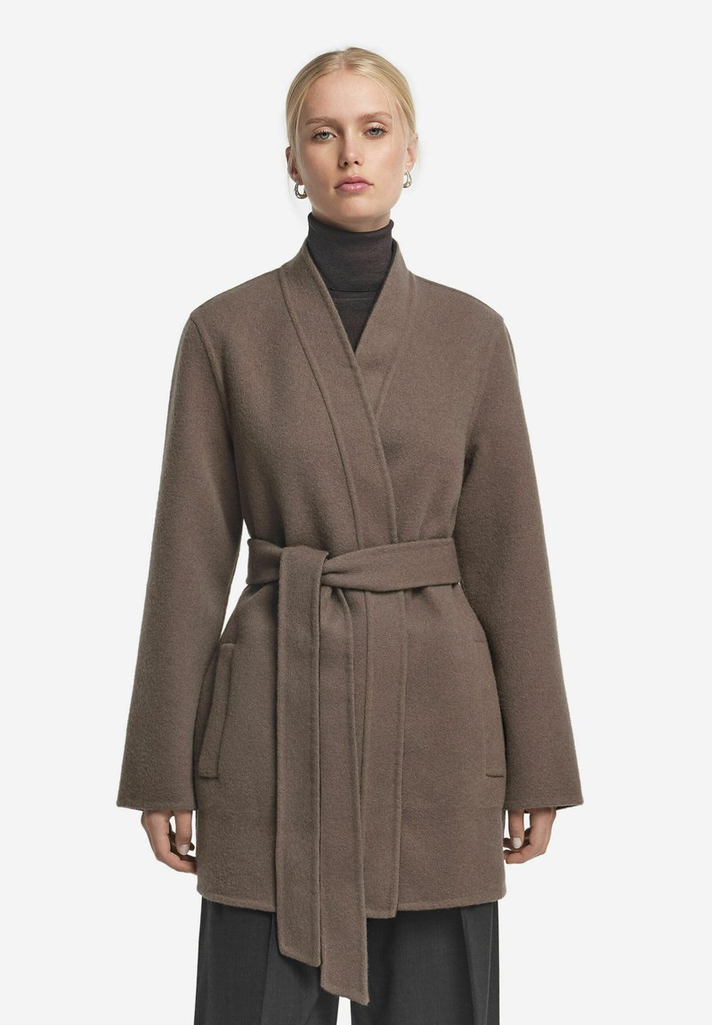 Brun svepjacka med långa ärmar, tillverkad av mjukt tyg, har ett bälte och sidofickor. Bärs över en svart polotröja.