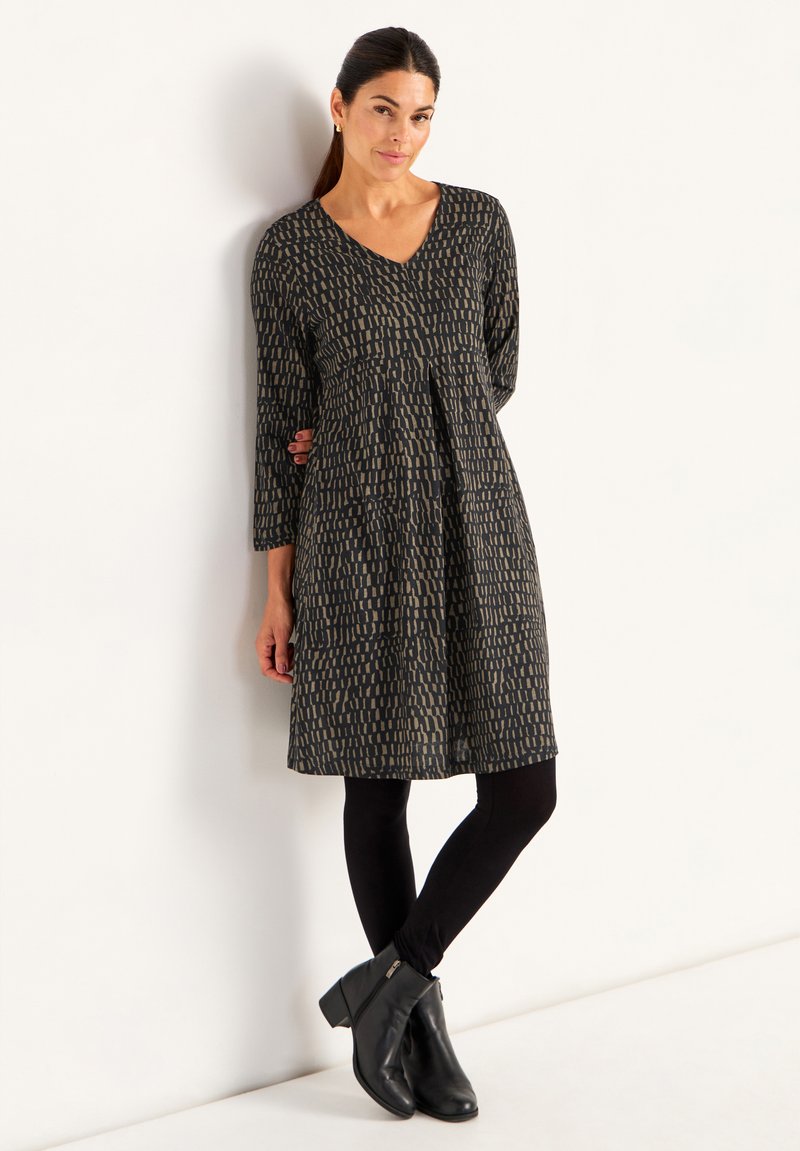 Robe noire et dorée à motifs avec un décolleté en V, des manches trois-quarts et une silhouette évasée, associée à des leggings noirs et des bottines.
