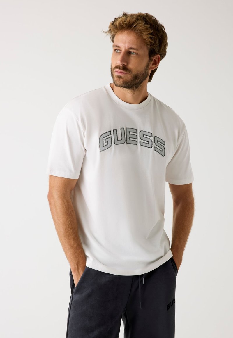 T-shirt bianco a maniche corte con il logo "GUESS" in grigio scuro al centro del petto. Il modello indossa pantaloni grigio scuro con dettagli del logo. Vestibilità casual.