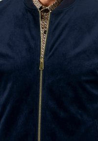 Granatowa kurtka bomberka wykonana z miękkiego materiału przypominającego zamsz, z złotym zamkiem błyskawicznym i ozdobnym kołnierzem z wzorem. Gładka faktura, klasyczny design.