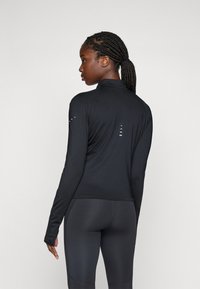 Femme avec une queue de cheval tressée, portant un haut de sport noir à manches longues ajusté et des legging, se tenant de côté contre un fond uni.