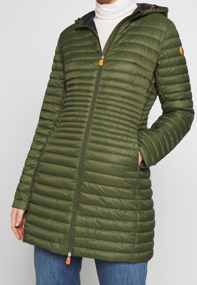 Veste matelassée vert olive avec un matelassage horizontal, des manches longues, une capuche et une fermeture éclair à l'avant. Équipée d'une tirette de fermeture éclair orange et de poches.