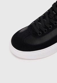 Black sneaker featuring a blend of smooth leather and suede upper, round toe, thick white rubber sole, and black laces.