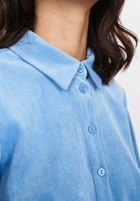 Camicia di velluto a coste azzurro chiaro con colletto appuntito, caratterizzata da bottoni blu coordinati e una texture morbida, vista da una prospettiva da spalla in su.