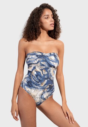 Žena s kovrčavom kosom, odjevena u kupaći kostim bez naramenica plave, bijele i bež uzorka, stoji ispred jednostavne svijetle pozadine.