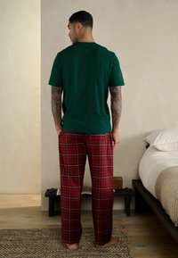 Maglietta verde in cotone abbinata a pantaloni da pigiama a quadri rossi. Presenta un collo tondo e una vestibilità comoda. Tessuto liscio con tatuaggi visibili sulle braccia.