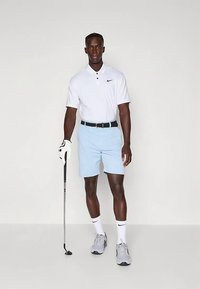 Vit polo-shirt med en svart Nike-logga, ljusblå shorts, svart bälte, golfhandske och grå sportskor, stående med en golfklubba.