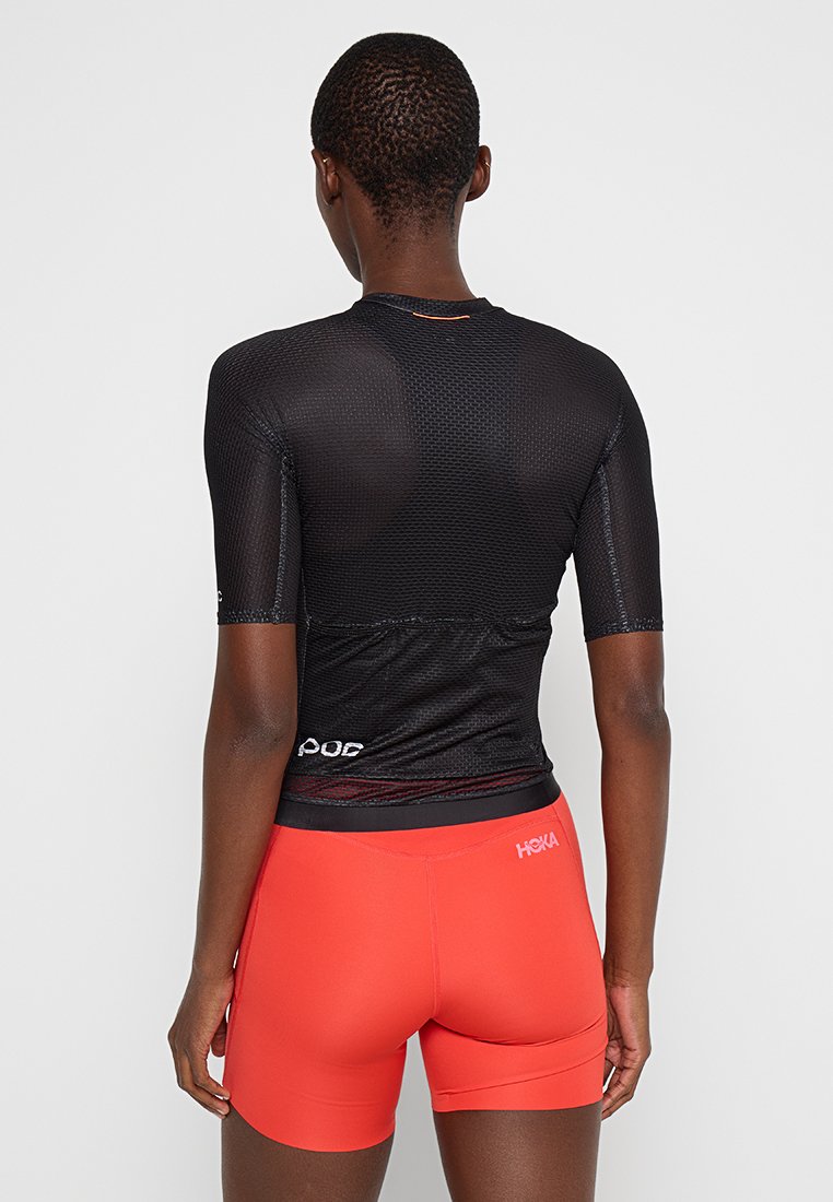 Schwarzes, kurzärmeliges Sportoberteil mit Netzstruktur und Logo auf dem Rücken. Kombiniert mit knallroten, figurbetonten Shorts.