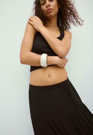 Young woman with curly hair wearing a black crop top and skirt, holding her arms crossed with a large white bracelet on her wrist.