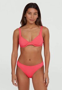 Kvinna med långt mörkt hår som bär en korallrosa bikini med en stödjande bygelöverdel och matchande underdelar, stående mot en ljus neutral bakgrund.