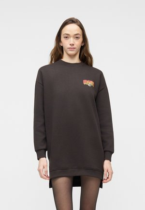 Frau trägt ein dunkelbraunes, übergroßes Sweatshirtkleid mit einer kleinen, bunten Grafik auf der linken Brust, kombiniert mit schwarzen transparenten Strumpfhosen.