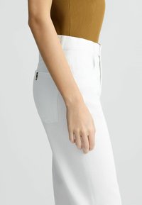 Pantalones blancos de talle alto con cremallera visible. El tejido parece tener una ligera textura. El bolsillo lateral presenta un pequeño acento dorado.