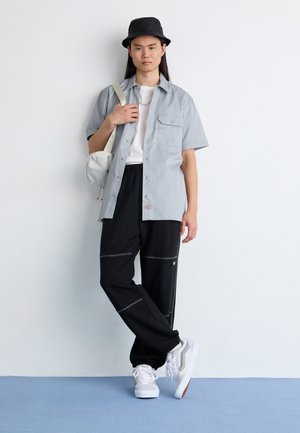 Camicia grigia chiara a maniche corte, pantaloni sportivi neri con cuciture a contrasto, sneaker bianche e un cappello a secchiello nero. Borsa a spalla piccola attaccata.
