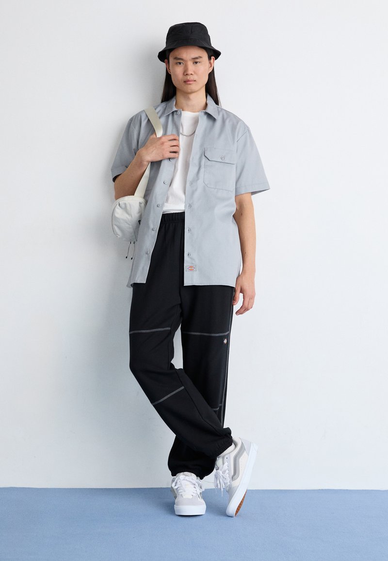 Helles graues Kurzarmshirt, schwarze Sweatpants mit Kontrastnähten, weiße Sneakers und ein schwarzer Bucket Hat. Kleine Umhängetasche befestigt.