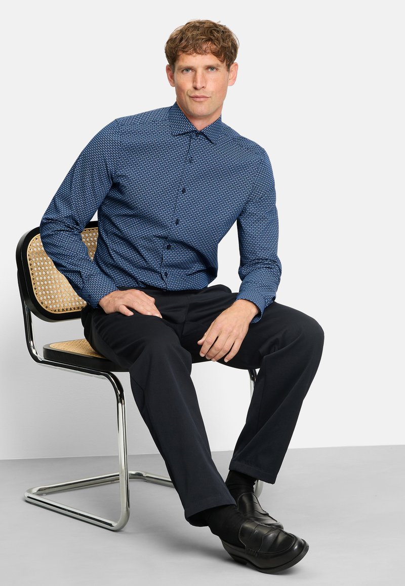 Man sitting on a modern chair wearing a blue patterned long-sleeve shirt, black pants, black socks, and black leather shoes.