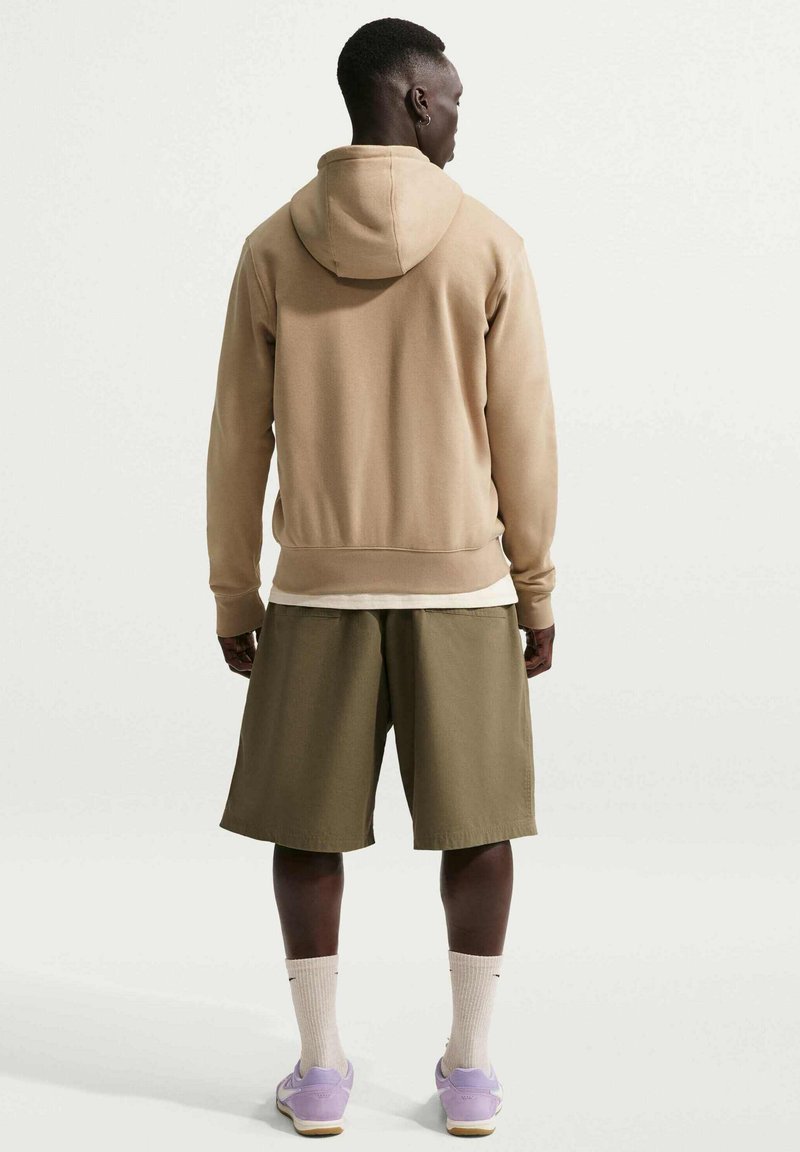 Man standing with back facing camera, wearing tan hoodie, olive green shorts, white socks, and light purple sneakers against plain background.