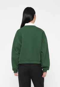 Felpa verde corta con maniche lunghe, realizzata in tessuto morbido, presenta un colletto a coste e una vestibilità rilassata, indossata sopra pantaloni neri.