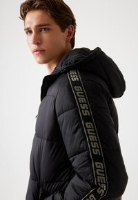 Chaqueta de plumón negra con capucha y cremallera lateral, con detalles de marca "GUESS" en beige a lo largo de las mangas; material suave y texturizado.