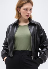 Νεαρή γυναίκα με κοντά μαλλιά που φοράει χρυσούς κρίκους αυτιών, μαύρο δερμάτινο μπουφάν με φερμουάρ, χακί πράσινο πουκάμισο και μαύρο παντελόνι.