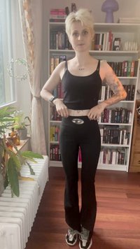 Fekete ujjatlan felső és fekete trapéz fazonú leggings, nagy logóval a deréknál. A modell egy könyvespolcaival rendelkező szobában áll.