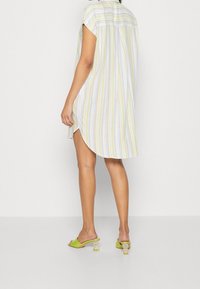 Gestreiftes Hemdkleid in Pastellfarben, mit kurzen Ärmeln, seitlichen Schlitzen und leichtem Stoff. Getragen mit grünen Rutschsandalen mit klaren Blockabsätzen.