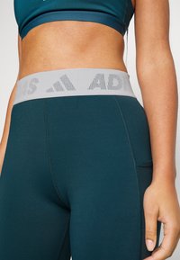 Turkoosi urheiluhousut, joissa on leveä harmaa elastinen vyötärö, jossa on painettu logo. Sujuva pinta ja tikkaus yksityiskohdissa sivuilla.