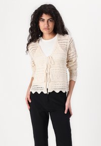 Pletený krémový cardigan s vlnkovaným lemem, otevřenými vzory a zavazováním vpředu. Stylizovaný přes přiléhavé bílé tričko a černé kalhoty.