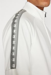 Giacca bianca con zip, caratterizzata da una striscia laterale grigia con dettagli del logo ripetuti. Tessuto morbido, vestibilità atletica e design colletto alto.
