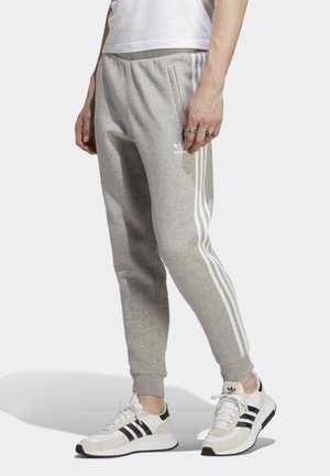 Persoon draagt grijze Adidas sweatpants met witte strepen en witte Adidas sneakers met zwarte strepen, staand op een witte achtergrond.