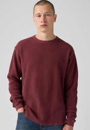Jeune homme aux cheveux courts portant un pull bordeaux texturé à manches longues et un jean bleu, une main dans la poche, debout devant un fond uni.