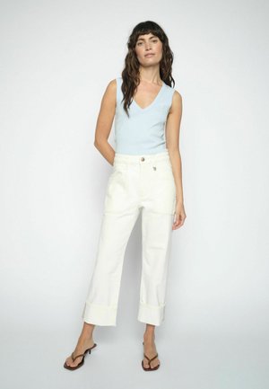 Hosszú, sötét hajú nő világoskék, ujjatlan V-nyakú felsőt, magasderekú, fehér, bőszárú nadrágot és barna magas sarkú szandált visel, áll.