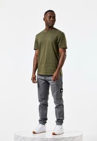 Un tricou verde măsliniu cu guler rotund și un mic logo negru, asortat cu pantaloni cargo gri, prevăzuți cu buzunare laterale și o pată neagră, și adidași albi.