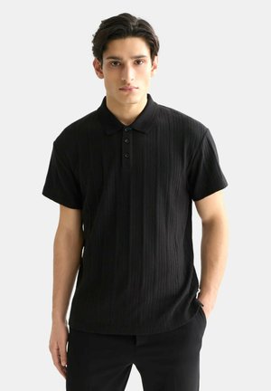 Junger Mann mit dunklen Haaren, der ein schwarzes, kurzärmeliges, strukturiertes Poloshirt und schwarze Hose vor einem schlichten hellen Hintergrund trägt.