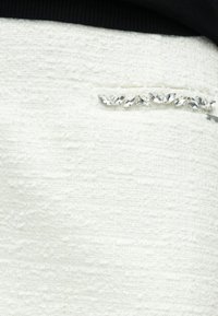 Weißer, strukturierter Stoff mit leichtem Glanz, der nahe der Naht eine Reihe klarer, birnenförmiger Edelsteine als Akzent aufweist.