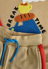 Tasse jaune avec de la vapeur et un pain sur une théière bleue brodée sur un tissu beige avec le texte "BREAK TIME" et un pantalon beige avec un cordon bleu.