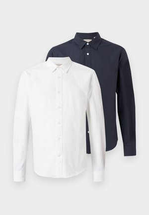 Duas camisas de manga longa, uma branca à frente e uma azul-marinho atrás, exibidas sobre um fundo claro e simples.