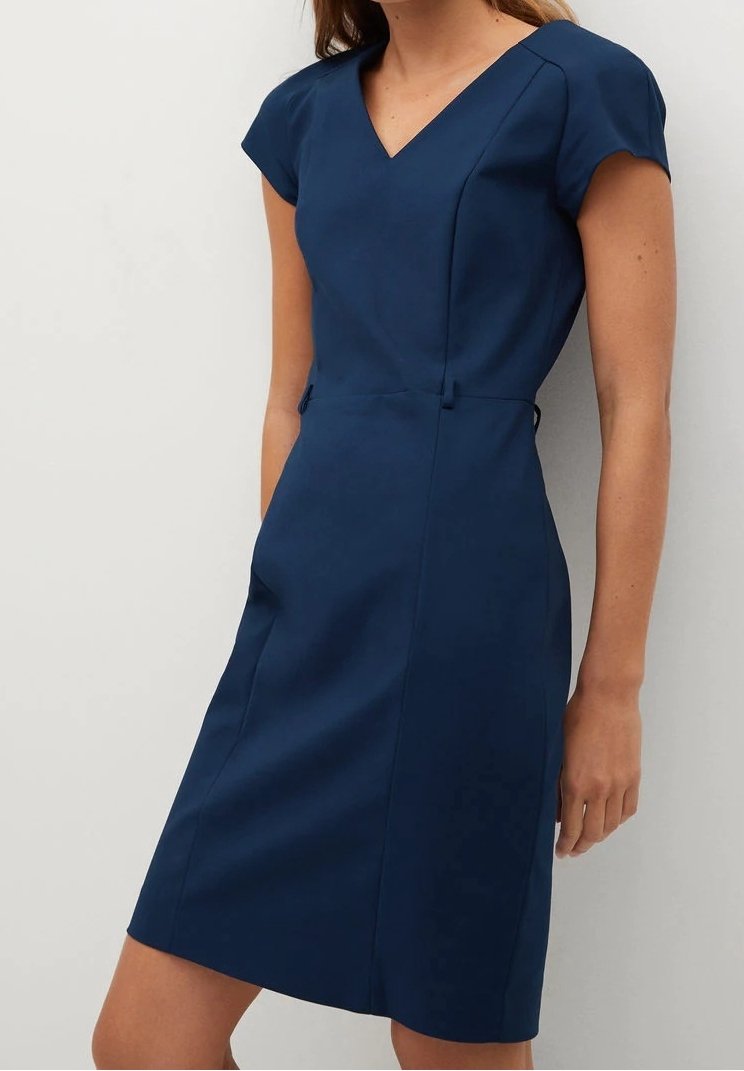 Femme portant une robe bleu marine ajustée, longueur genoux, à manches courtes et encolure en V, debout devant un fond clair uni.