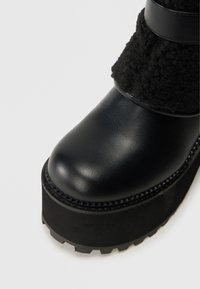 Tuvo plāna skats uz melnu ādas platformu zābaku ar biezu gumijas zoli un melnu vilnas maliņu ar ādas siksniņu detaļu.