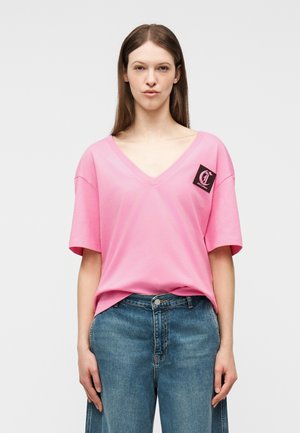 Młoda kobieta ubrana w luźny różowy T-shirt z dekoltem w serek, z małą czarną naszywką z logo na piersi, oraz w niebieskie dżinsy z wysokim stanem.