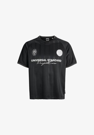 Sort kortærmet trøje lavet af glat stof. Har lodrette striber, logo-patches og fed hvid tekst, der står "UNIVERSAL STANDARD."