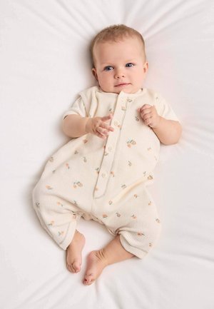 Bambino con occhi azzurri disteso su una coperta bianca, indossa un completo color crema con una piccola stampa floreale pesca, che tende una mano in avanti.