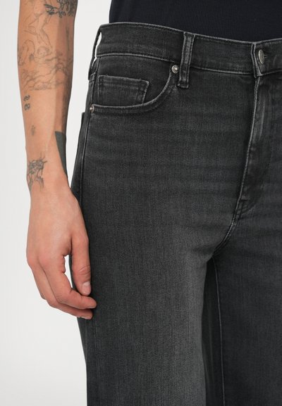 Mustad teksapüksid on kitsama lõikega, keskmise kõrgusega vöökohaga, kontrastsete õmblustega ja viie taskuga, kujutatud tindi tätoveeringutega käel.