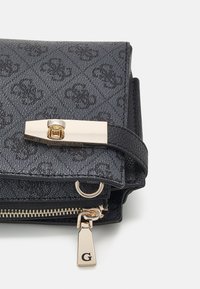 Svart texturerad handväska med ett guldspänne, dragkedjedetalj och en charm med bokstaven "G." Designen är stilren med en kompakt form.
