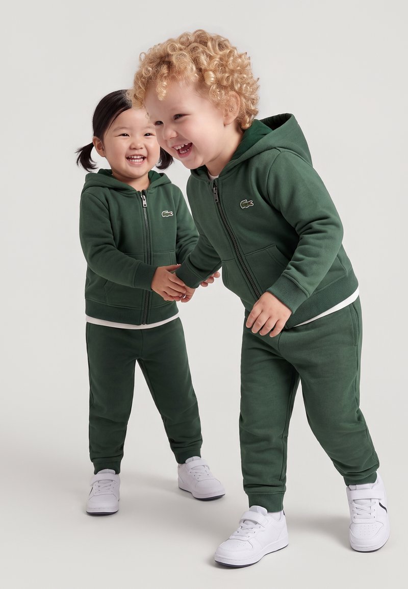 Deux jeunes enfants portant des survêtements à capuche vert foncé assortis et des baskets blanches, souriants et se tenant la main.